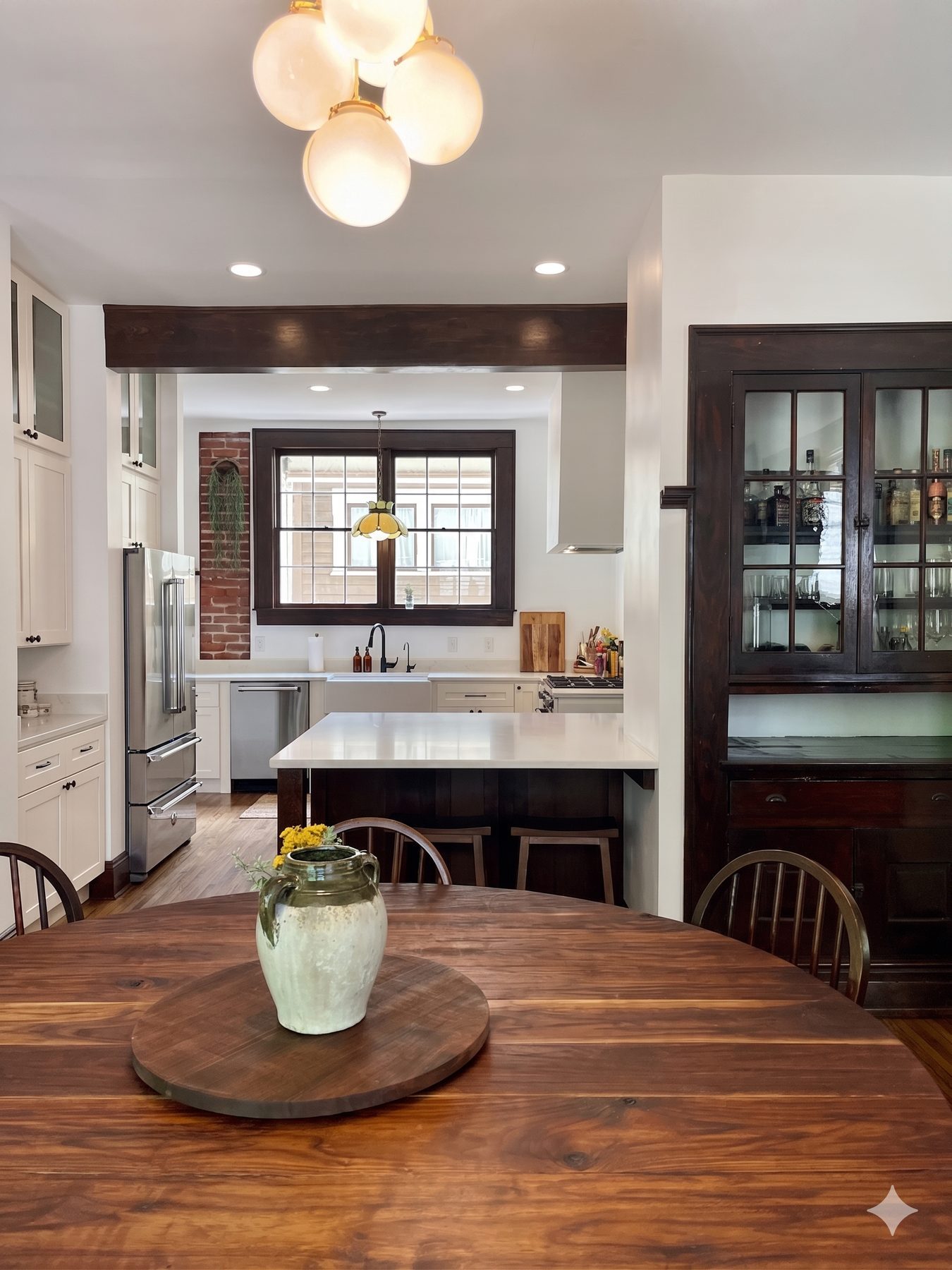 Birmingham kitchen — view from round wood dining table through to the renovated kitchen with globe pendant, dark beam, glass cabinets, and brick chimney beyond
